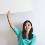 Real-Time Marketing - Cheerful Asian woman sitting cross legged on floor against white wall in empty apartment and showing white blank banner