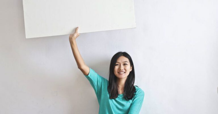 Real-Time Marketing - Cheerful Asian woman sitting cross legged on floor against white wall in empty apartment and showing white blank banner