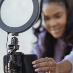 Tutorials - Cheerful young African American female blogger in stylish sweater smiling while setting up camera of smartphone attached to tripod with ring light before recording vlog
