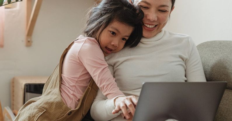 User-Generated Content - Cheerful smiling Asian woman browsing modern netbook while hugging with cute content daughter on comfy sofa in cozy living room