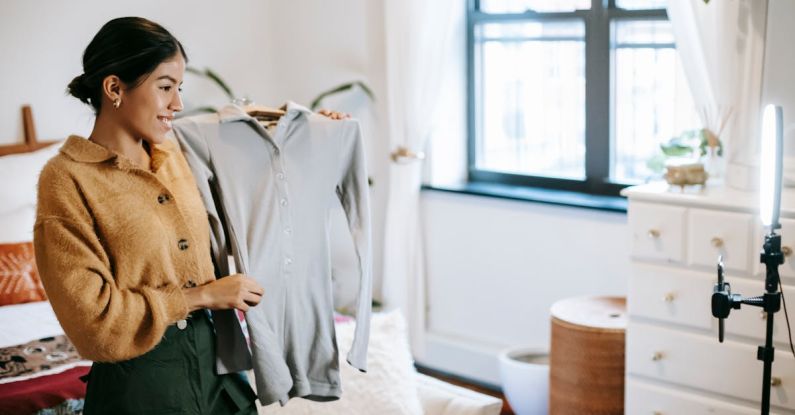 Social Media Promotion - Positive ethnic female blogger demonstrating jacket on hanger during live stream on mobile phone placed on shining lamp