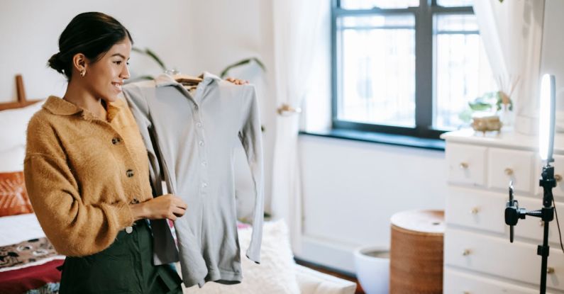 Social Media Promotion - Positive ethnic female blogger demonstrating jacket on hanger during live stream on mobile phone placed on shining lamp